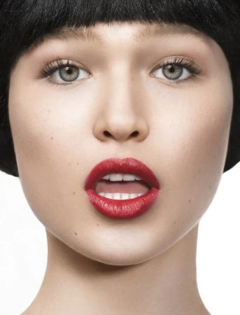 Close-up portrait of a woman with bold red lipstick, dark hair with bangs, and pale skin against a white background.