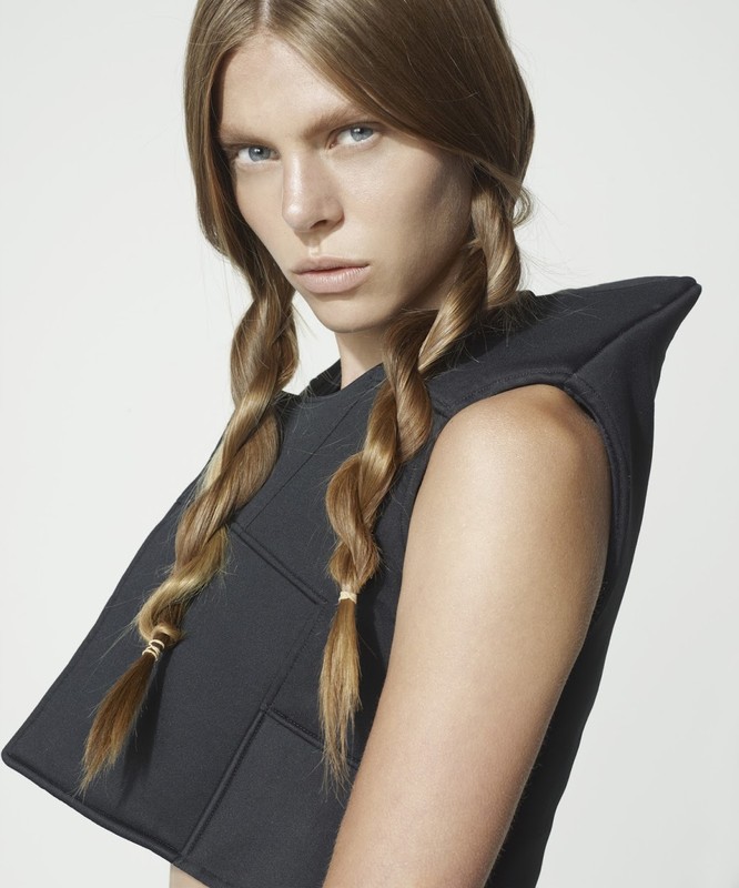 Portrait of a woman with blonde hair styled in braids, wearing a geometric black top against a neutral studio backdrop.