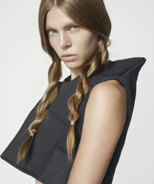 Portrait of a woman with blonde hair styled in braids, wearing a geometric black top against a neutral studio backdrop.