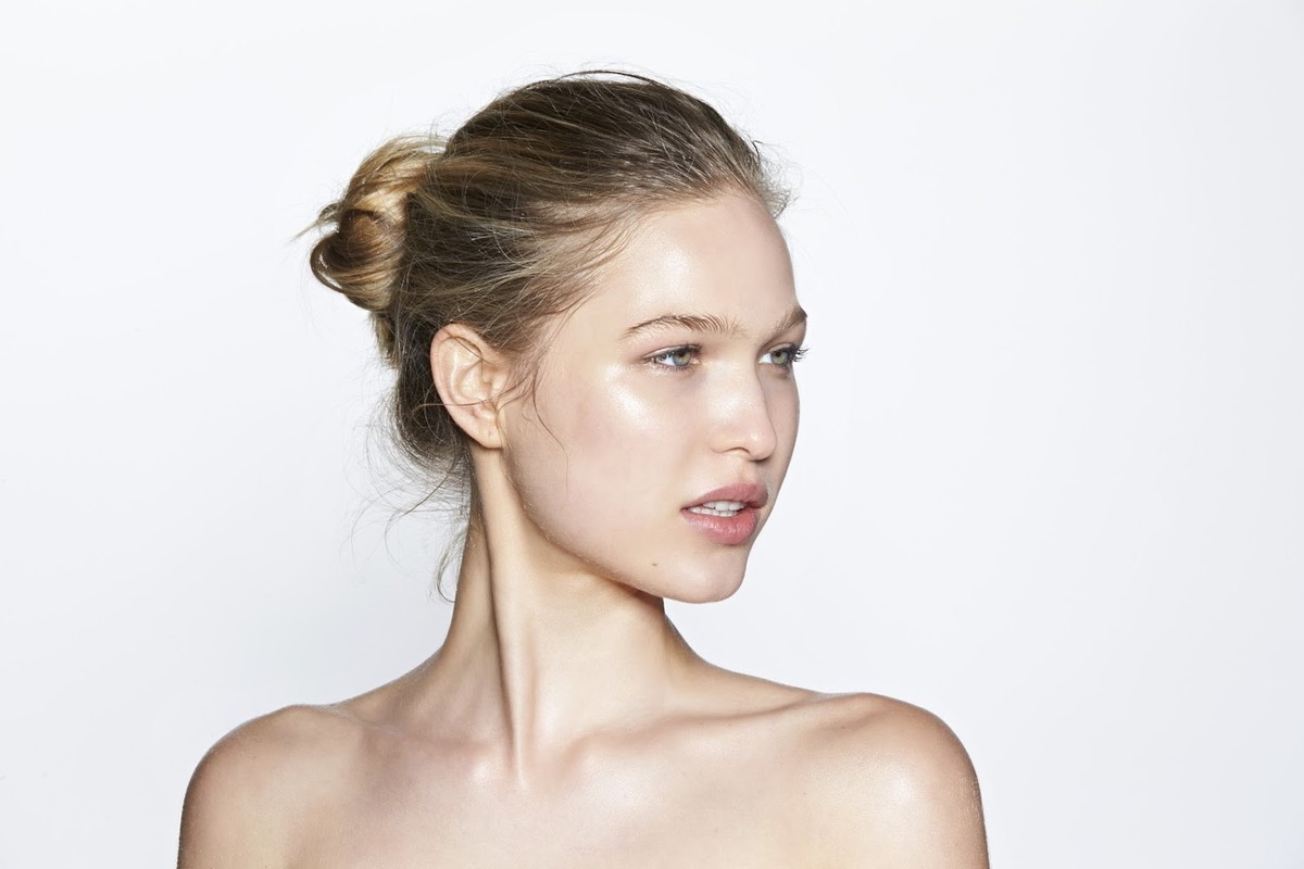 Close-up portrait of a fair-skinned young woman with blonde hair styled in a bun, posing against a bright white backdrop in a beauty shot.