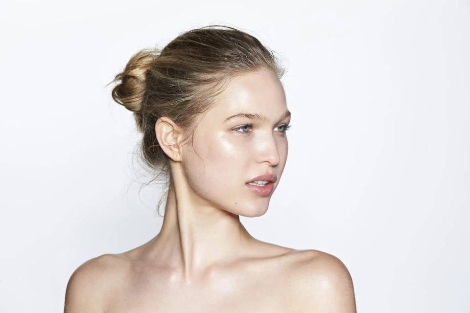 Close-up portrait of a fair-skinned young woman with blonde hair styled in a bun, posing against a bright white backdrop in a beauty shot.