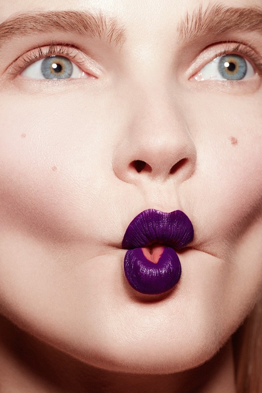 Close-up portrait of a young fair-skinned woman with blue eyes making a fish face with purple lipstick covering her lips.
