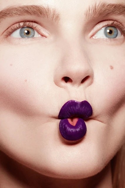 Close-up portrait of a young fair-skinned woman with blue eyes making a fish face with purple lipstick covering her lips.