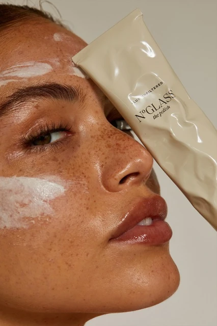 Close-up of a woman's face with a beauty product tube covering part of her face, featuring freckles, glossy lips, and smooth skin.