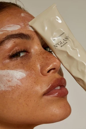 Close-up of a woman's face with a beauty product tube covering part of her face, featuring freckles, glossy lips, and smooth skin.