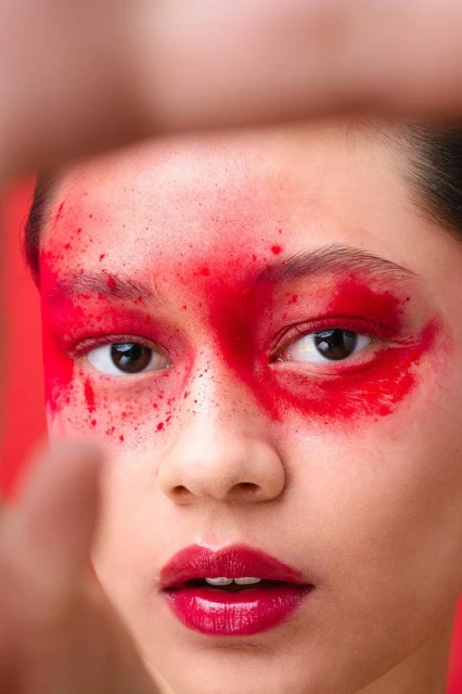 Close-up portrait of an adult female with striking red face paint and matching red lipstick against a vivid red backdrop, looking at the camera.