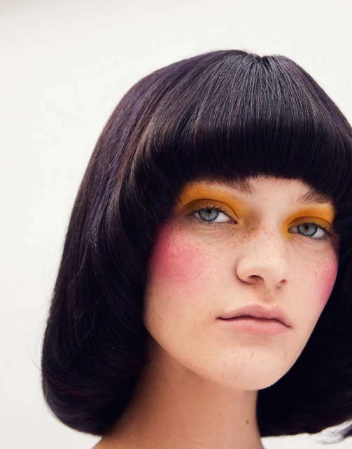 Close-up portrait of an adult female with short dark hair, yellow and pink makeup, and freckles, against a plain white background.