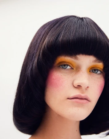 Close-up portrait of an adult female with short dark hair, yellow and pink makeup, and freckles, against a plain white background.