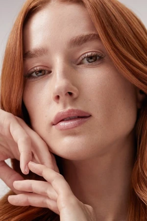 Close-up portrait of a woman with red hair and light makeup holding her hand near her chin against a neutral background.