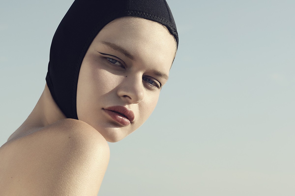Portrait of a young woman with neutral expression wearing a black swim cap and dark winged eyeliner against a light blue background.
