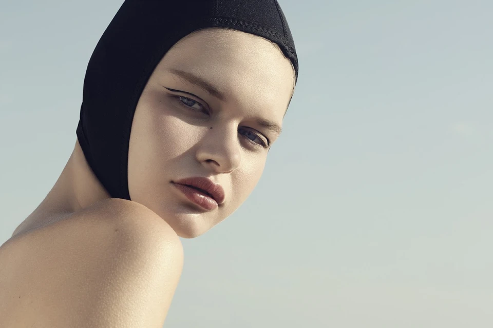 Portrait of a young woman with neutral expression wearing a black swim cap and dark winged eyeliner against a light blue background.