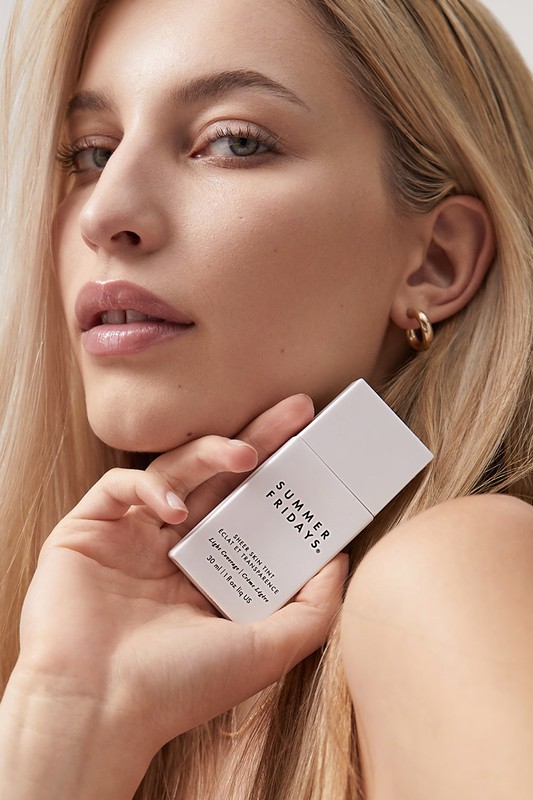 Blonde woman holds Summer Fridays skincare, showcasing its packaging against a neutral background for a beauty and fashion theme.