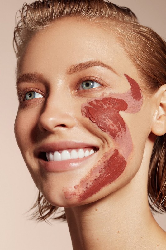 Close-up studio shot of a fair-skinned woman with blonde hair, smiling with a skincare product applied as a face mask.