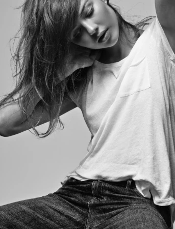 Monochrome portrait of a woman with tousled hair wearing a white t-shirt and dark jeans against a light background
