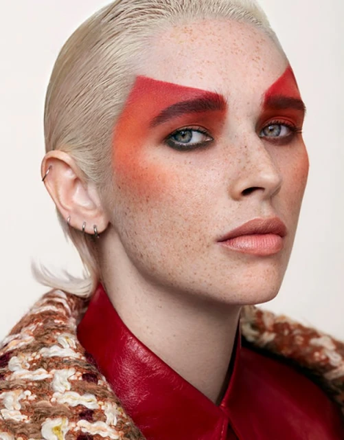 Portrait of a woman with striking red makeup, blonde hair, wearing a patterned jacket and red leather shirt against a white background.