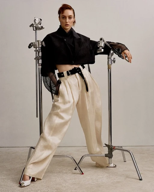 Fashionable woman in a black crop top and beige pants strikes a pose in a studio setting, leaning on a metal c-stand, showcasing modern fashion.