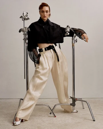 Fashionable woman in a black crop top and beige pants strikes a pose in a studio setting, leaning on a metal c-stand, showcasing modern fashion.