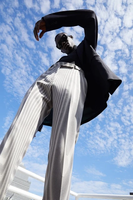 Low-angle view of an African-American male model wearing a dark blazer and patterned pants against a cloudy blue sky. Fashion shot.