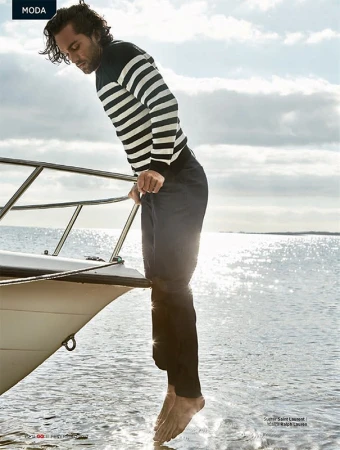 Adult male model on a boat wearing striped sweater and dark pants steps barefoot into water with sunny sky background for a fashion editorial.