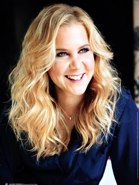 Smiling woman with wavy blonde hair and blue eyes wearing a navy blouse and delicate gold necklace, portrait shot.