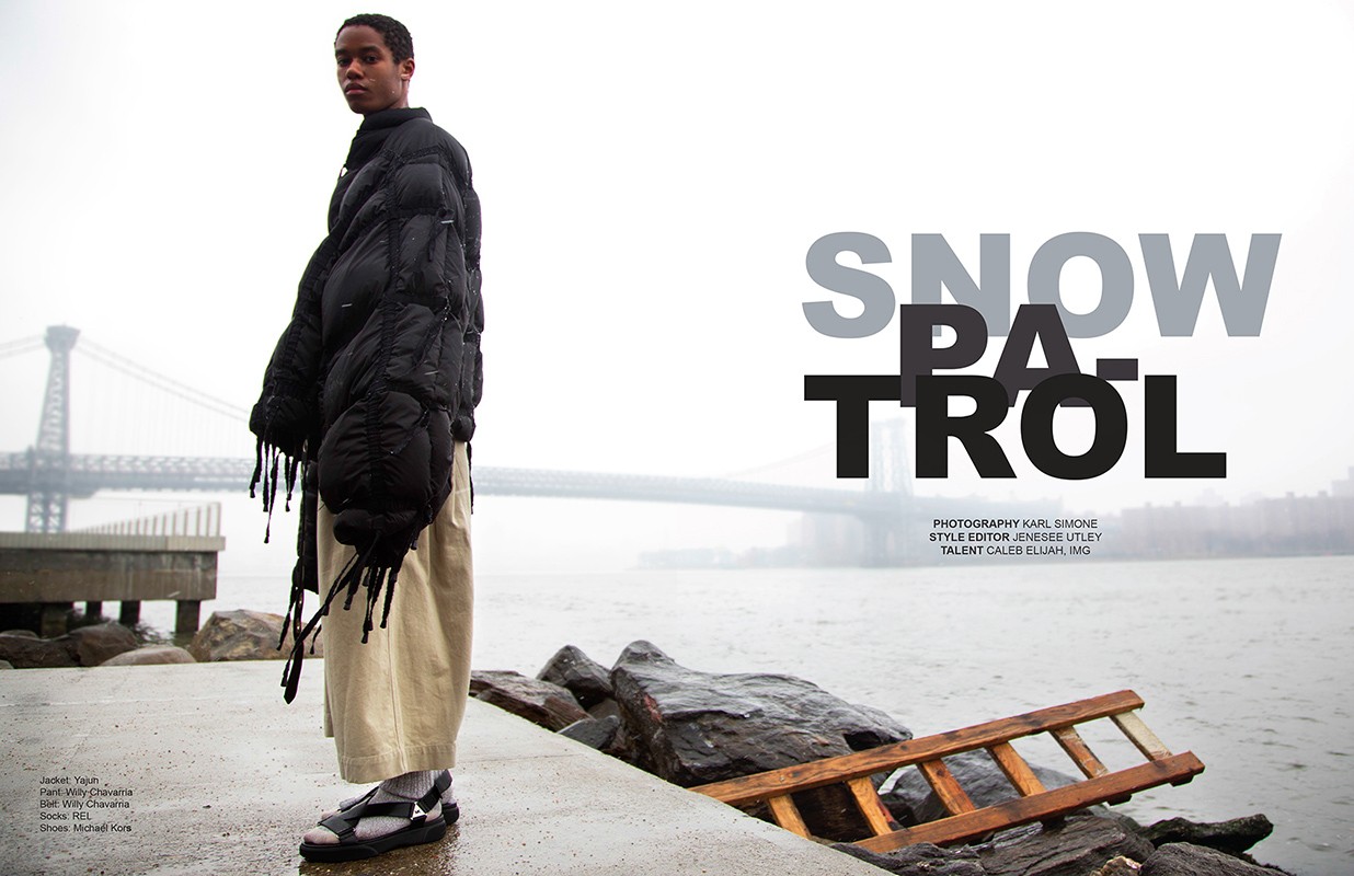 Fashion shot of a Black male model wearing a puffer jacket and sandals, standing on a waterside concrete dock near a bridge.