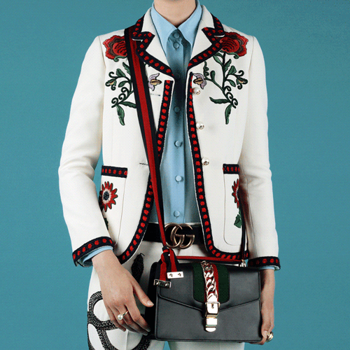 Fashionable person in floral embroidered jacket with leather handbag against a blue background in a studio setting, portrait view.