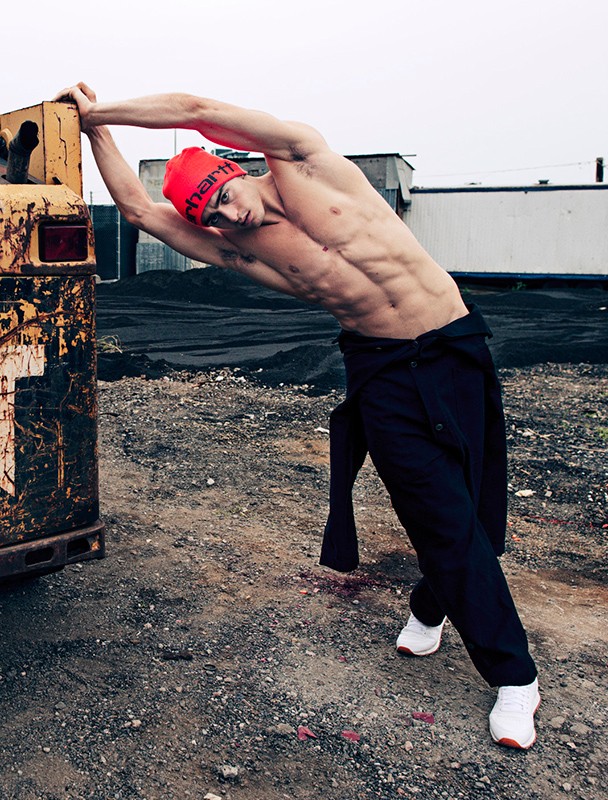 Athletic man in black pants and red beanie posing with a rusted yellow construction vehicle in an outdoor, industrial setting.