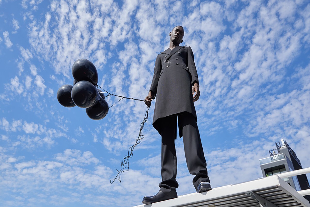 A tall, slender Black man stands on a rooftop holding a bunch of black balloons against a blue sky with scattered clouds, shot from a low angle.