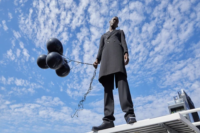 A tall, slender Black man stands on a rooftop holding a bunch of black balloons against a blue sky with scattered clouds, shot from a low angle.