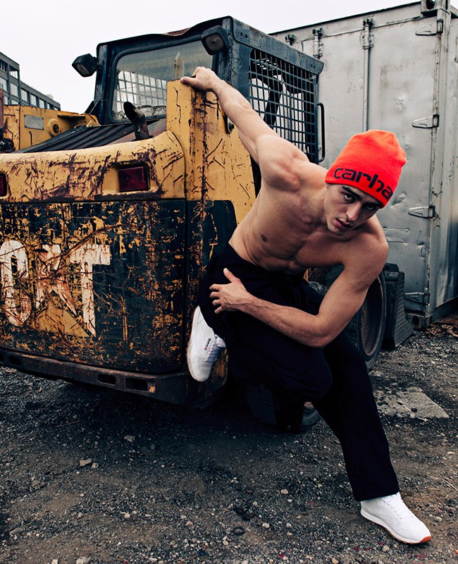 Male model in orange beanie poses with yellow CAT construction vehicle in outdoor shot against a neutral background in a gritty, urban setting.