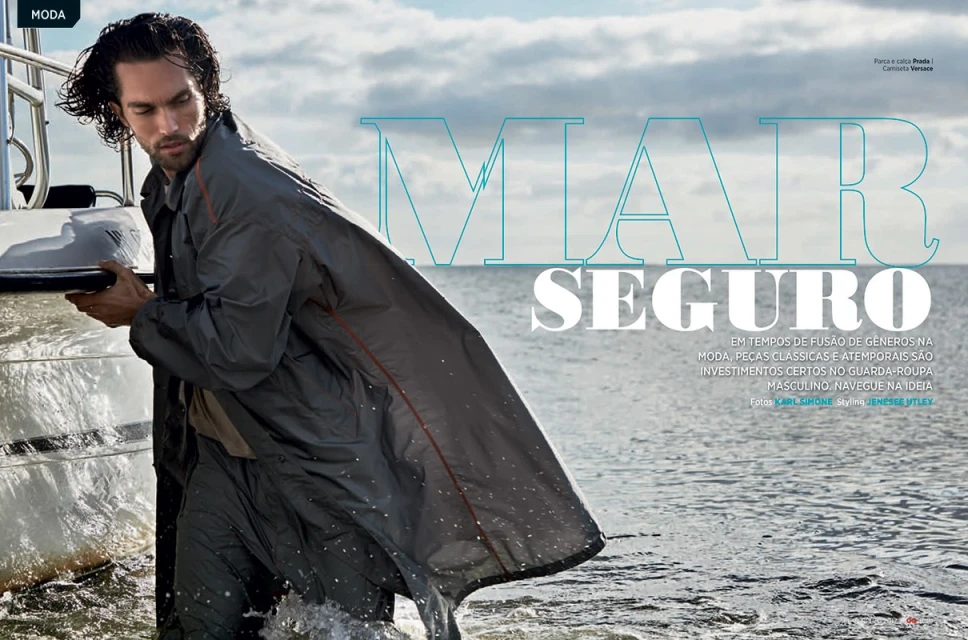 A man models gray outerwear while wading in the ocean next to a small boat, with cloudy sky and magazine text overlay.