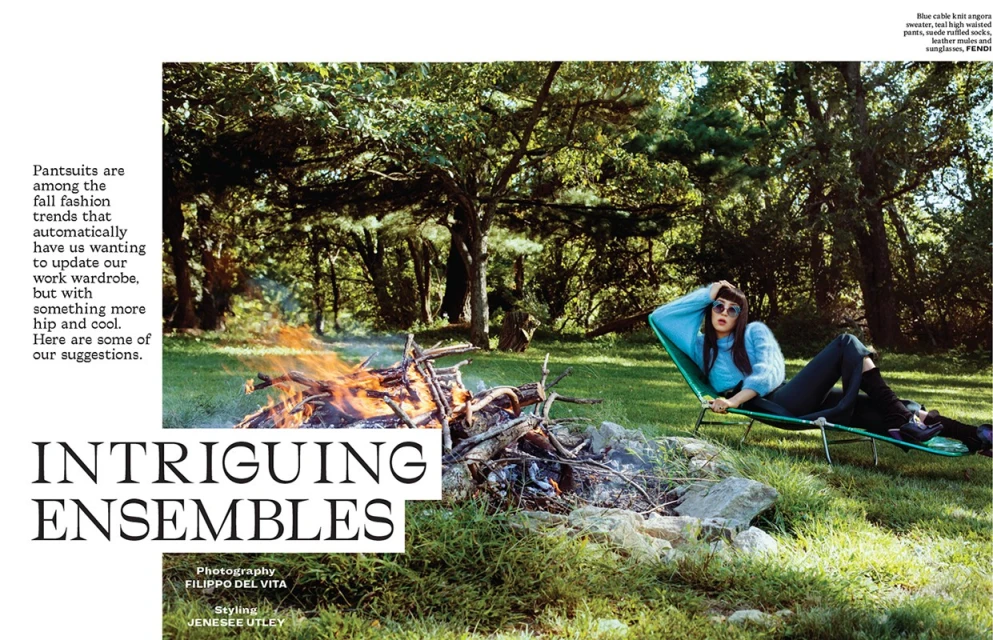 Fashion editorial shot outdoors of a woman in sunglasses lying in a lounge chair near a fire pit, wearing a blue sweater and dark pants.