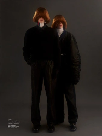 Two models with heavy red fringe covering their eyes wearing dark clothing against a plain grey background