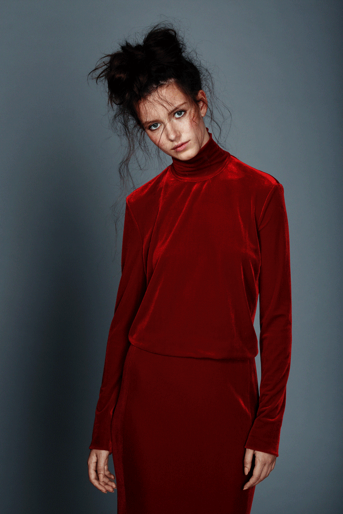 Portrait of a woman in a red velvet dress with a high neckline, posing against a grey background in a fashion style photograph.