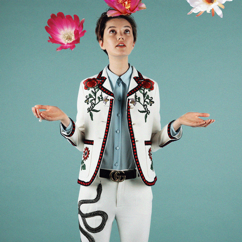 Woman in white suit with red floral embroidery posing with pink flowers floating around her against a teal background in a studio shot.