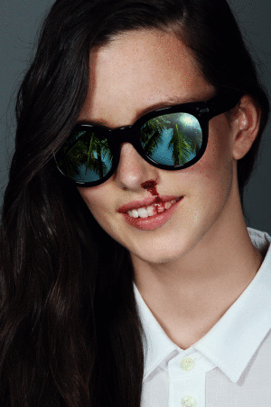 Portrait of a young woman with dark hair, black sunglasses reflecting palm trees, and a nosebleed, set against a grey backdrop.