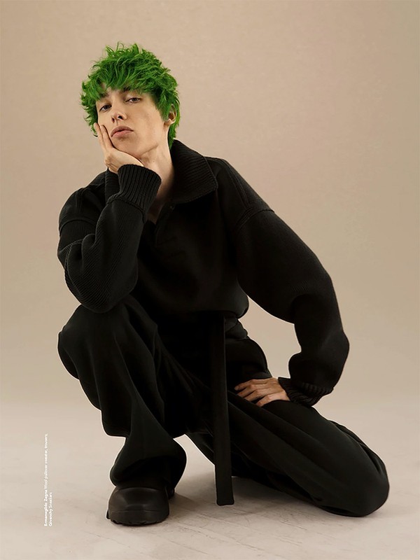 An individual with green hair poses in a photo studio. They are wearing a black sweater and matching pants, posed in front of a tan backdrop.