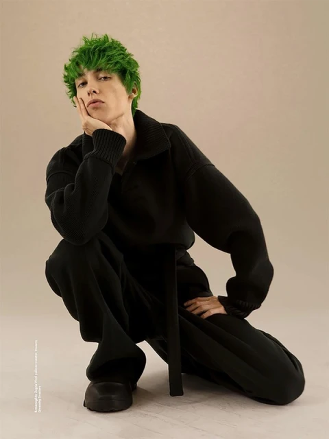 An individual with green hair poses in a photo studio. They are wearing a black sweater and matching pants, posed in front of a tan backdrop.