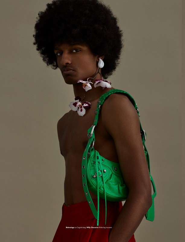 Fashion portrait of a Black male model with afro, wearing a green purse, flower necklace and earrings, and red pants against beige background.