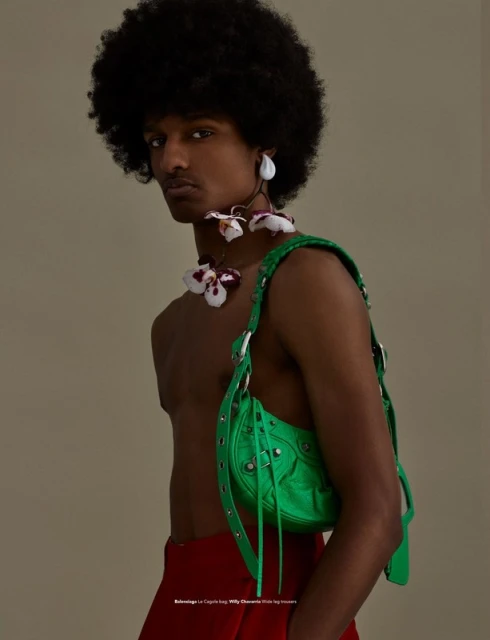 Fashion portrait of a Black male model with afro, wearing a green purse, flower necklace and earrings, and red pants against beige background.