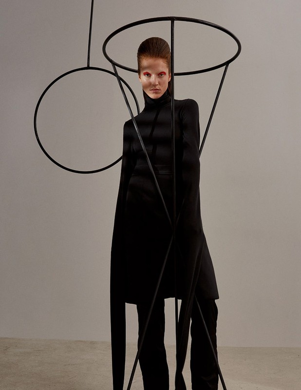 Fashionable woman in black turtleneck and pants poses indoors with dramatic makeup, standing among tall, abstract black metal forms.