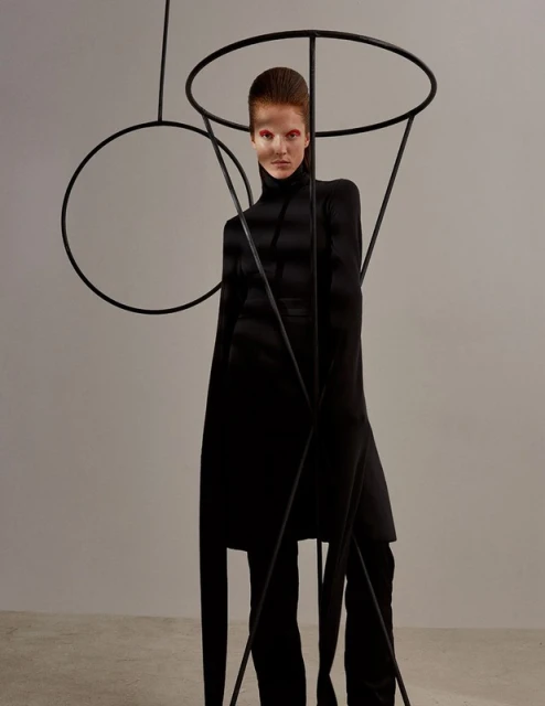 Fashionable woman in black turtleneck and pants poses indoors with dramatic makeup, standing among tall, abstract black metal forms.