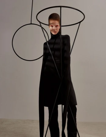 Fashionable woman in black turtleneck and pants poses indoors with dramatic makeup, standing among tall, abstract black metal forms.