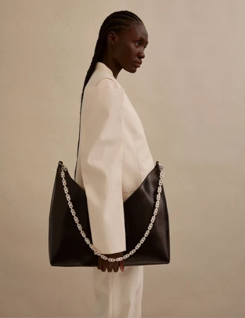 Black woman wearing a cream structured jacket holding a large black leather shoulder bag with a silver chain strap against a neutral wall.