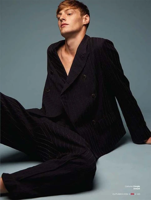 Blonde male model in black pinstripe suit posing indoors.  Fashion editorial photography of man in suit, sitting against solid background.