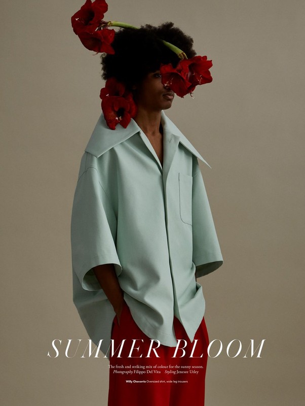 Fashion shot of a person with dark hair adorned with red flowers, wearing an oversized pale-green shirt and red pants on a neutral backdrop.