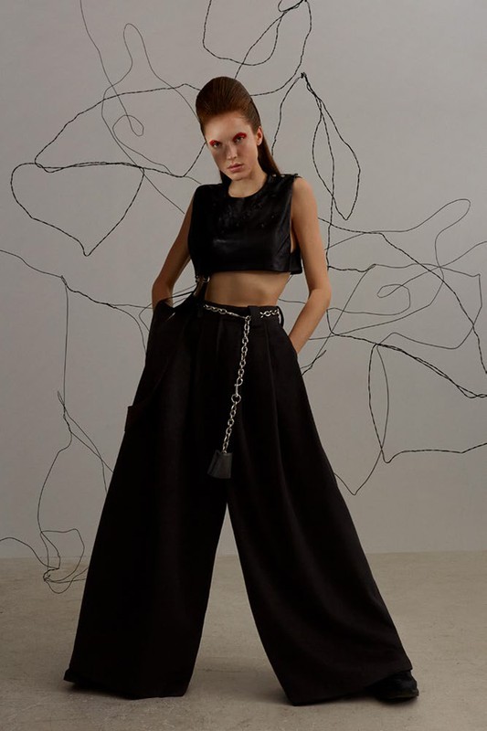A model poses in an editorial fashion shot, wearing a black crop top and wide-leg pants, accented by a chain belt in a photo studio.
