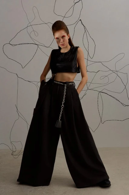 A model poses in an editorial fashion shot, wearing a black crop top and wide-leg pants, accented by a chain belt in a photo studio.