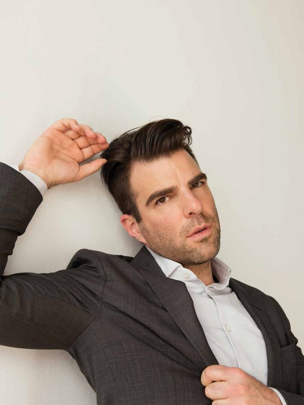 Portrait of an adult male in a grey suit and light shirt, leaning against a white wall while running a hand through his hair.