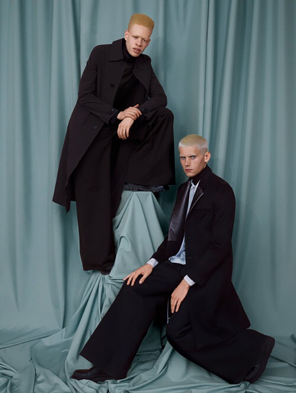 Two men with bleached blonde hair pose in black suits and wide-leg trousers, styled on a draped light-blue fabric-covered pedestal.